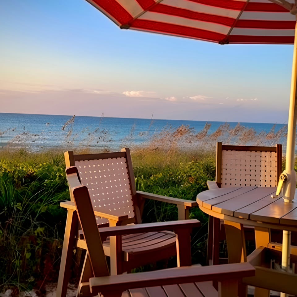 Outdoor dining area with chairs, table, and red-striped umbrella overlooking the ocean at sunset.