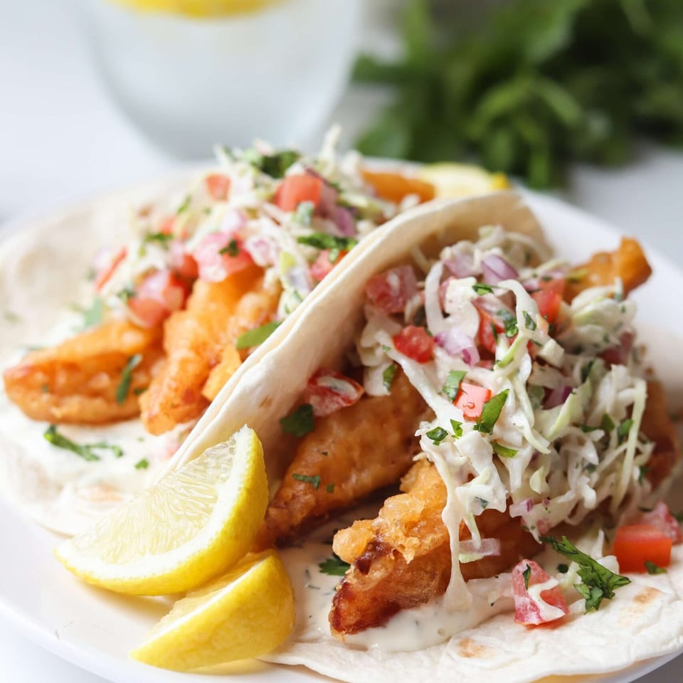 Two fish tacos with crispy fish, coleslaw, salsa, and lemon wedges on a white plate.