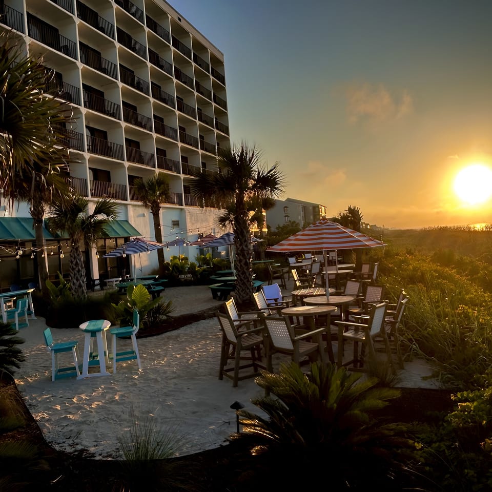 Sunset over a beachside restaurant patio with palm trees and hotel in the background.