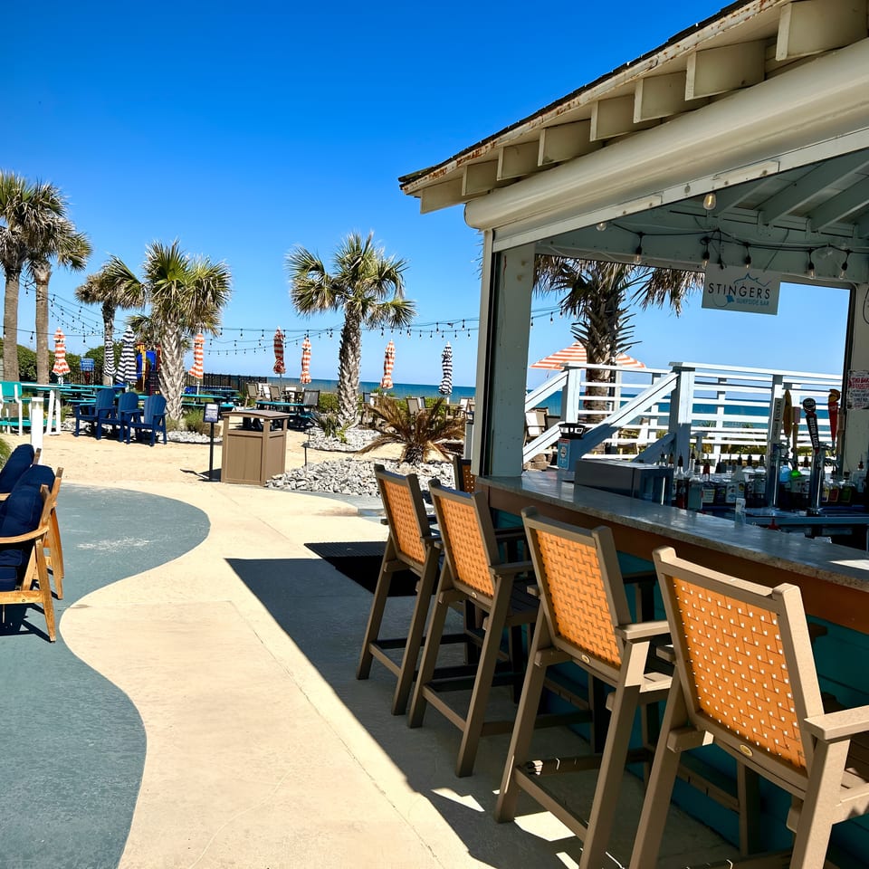 Outdoor bar with orange stools, palm trees, and ocean views under a clear blue sky.